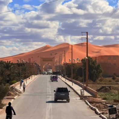 A street in Merzouga leads to towering Erg Chebbi sand dunes, capturing the vast desert landscape of southeastern Morocco.